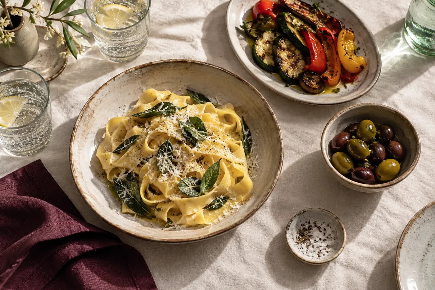 Handmade pasta, vegetables and olives arranged on an ivory tablecloth.